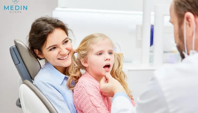 Uśmiechnięta mama towarzyszy córce podczas zabiegu lakowania zębów u dzieci. Dentysta bada jamę ustną dziewczynki.